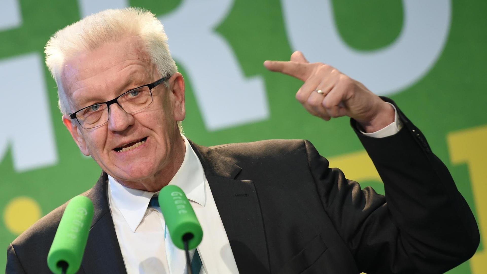 Sie sehen Baden-Württembergs Ministerpräsident Kretschmann, er spricht in zwei grüne Mikrofone.