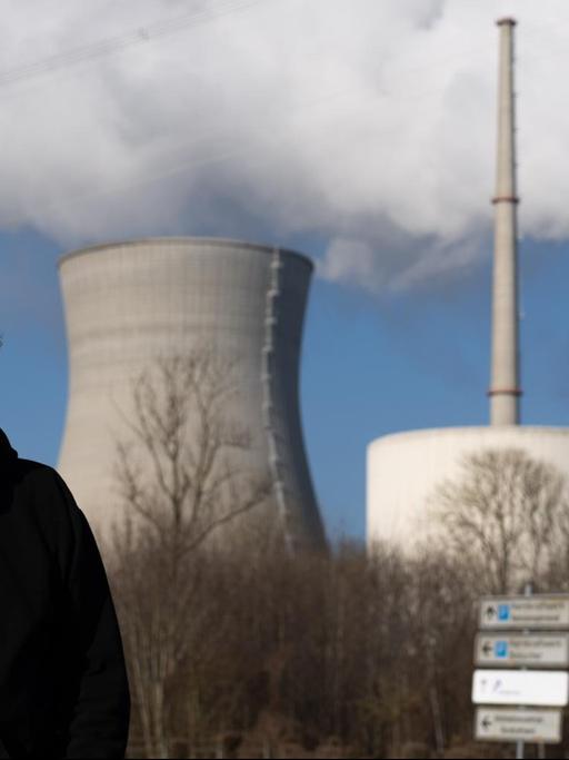 Ein Mann mit grauen Haaren und Schnauzbart steht im schwarzen Kapuzenshirt vor einem Atomkraftwerk, aus dem weißer Rauch in den blauen Himmel aufsteigt.