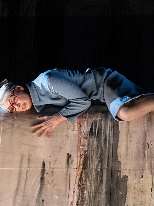Szenenbild aus der Oper "Toteis" von Manuela Kerer & Matthias Plattner mit Isabel Seebacher als "Viktoria", die bäuchlings vor einem schwarzen Hintergrund auf einer grauen Mauer liegt und einen Verband um den Kopf trägt.