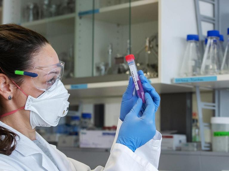Eine Wissenschaftlerin mit Mundschutz, Schutzbrille und Handschuhen hält ein Laborröhrchen mit der Aufschrift "SARS-CoV 2" in den Händen.