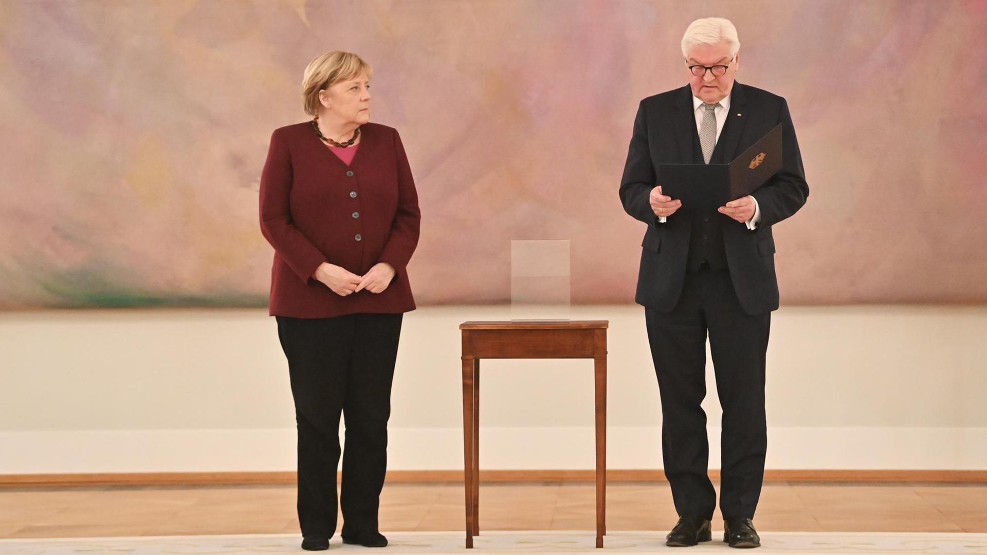 Bundespräsident Frank-Walter Steinmeier händigt Bundeskanzlerin Angela Merkel die Entlassungsurkunde aus.Beide stehen vor einem abstrakten rosa Gemälde, zwischen ihnen: ein hochbeiniger Beistelltisch.
