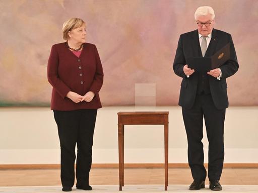 Bundespräsident Frank-Walter Steinmeier händigt Bundeskanzlerin Angela Merkel die Entlassungsurkunde aus.Beide stehen vor einem abstrakten rosa Gemälde, zwischen ihnen: ein hochbeiniger Beistelltisch.