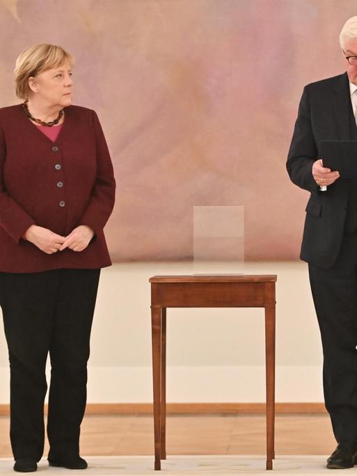 Bundespräsident Frank-Walter Steinmeier händigt Bundeskanzlerin Angela Merkel die Entlassungsurkunde aus.Beide stehen vor einem abstrakten rosa Gemälde, zwischen ihnen: ein hochbeiniger Beistelltisch.