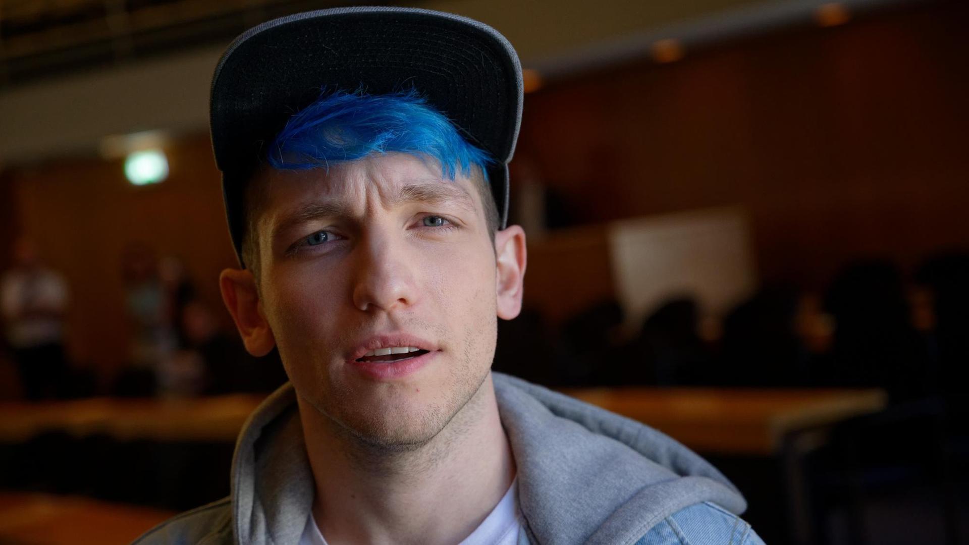 Youtuber Rezo, mit blau gefärbtem Haar und Basecap, schaut skeptisch in die Kamera, im Hintergrund: ein holzgetäfelter Veranstaltungssaal