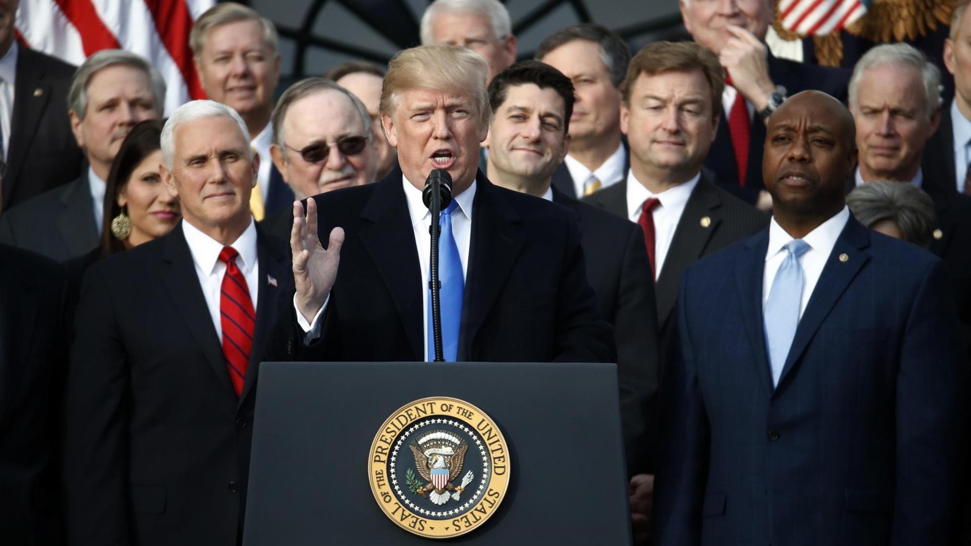Trump spricht gestikulierend an einem Rednerpult mit dem Präsidentenwappen, hinter ihm viele Republikaner. Trump spricht gestikulierend an einem Rednerpult mit dem Präsidentenwappen, hinter ihm viele Republikaner.