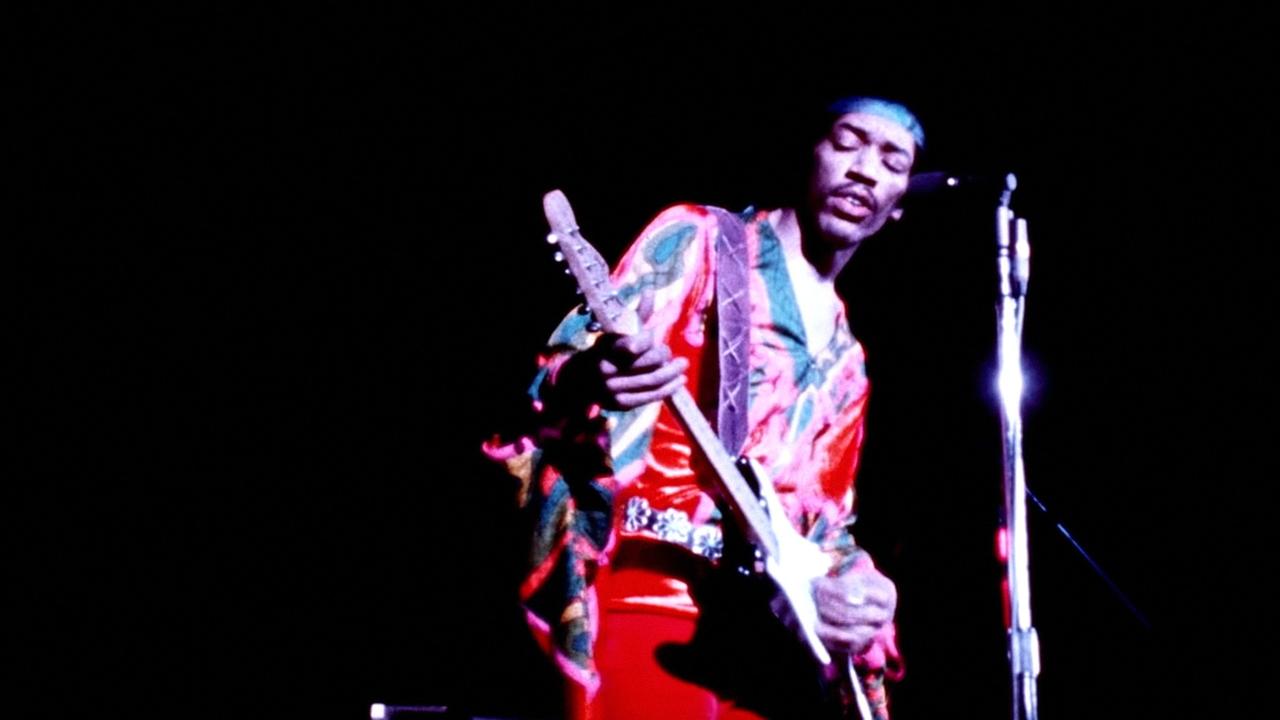 Jimi Hendrix steht mit Gitarre auf einer Bühne und trägt bunte Kleidung und ein Stirnband.