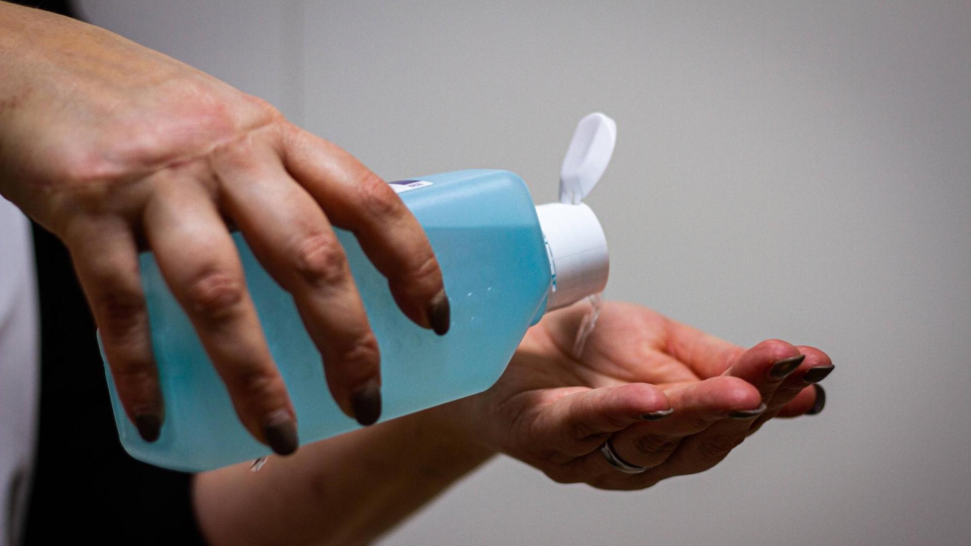 Eine Person hält eine große blaue Flasche Desinfektionsmittel in den Händen und kippt die Flüssigkeit in die linke Hand.