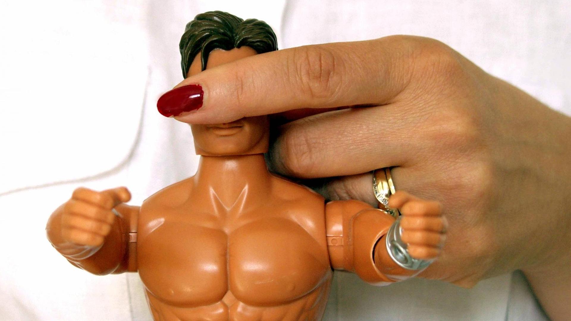 Frauenhand mit rot lackierten Fingernägeln halten der nackten Puppe Ken die Augen zu.
