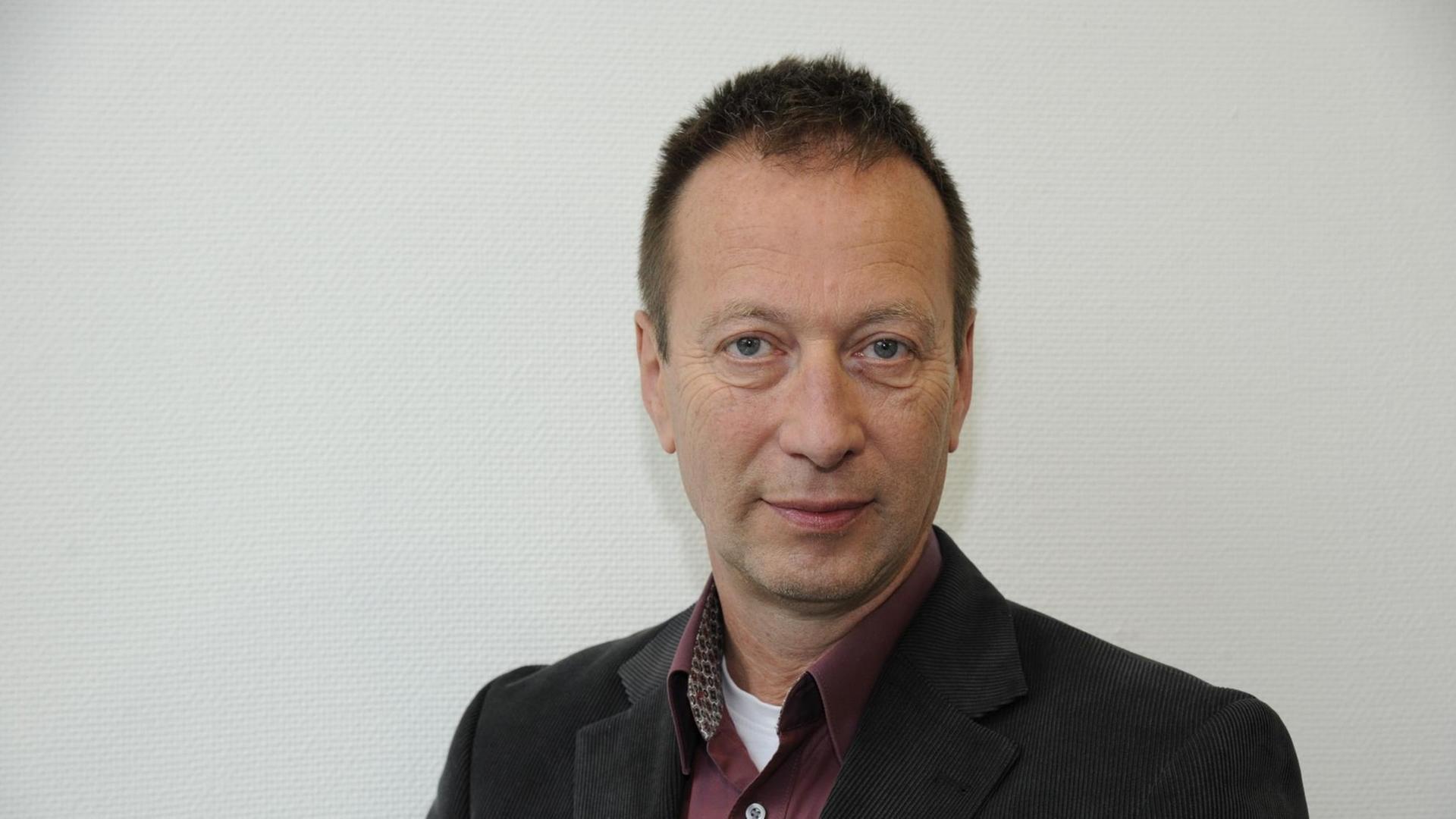 Der Schriftsteller und Übersetzer Hans Pleschinski steht lächelnd vor einer weißen Wand