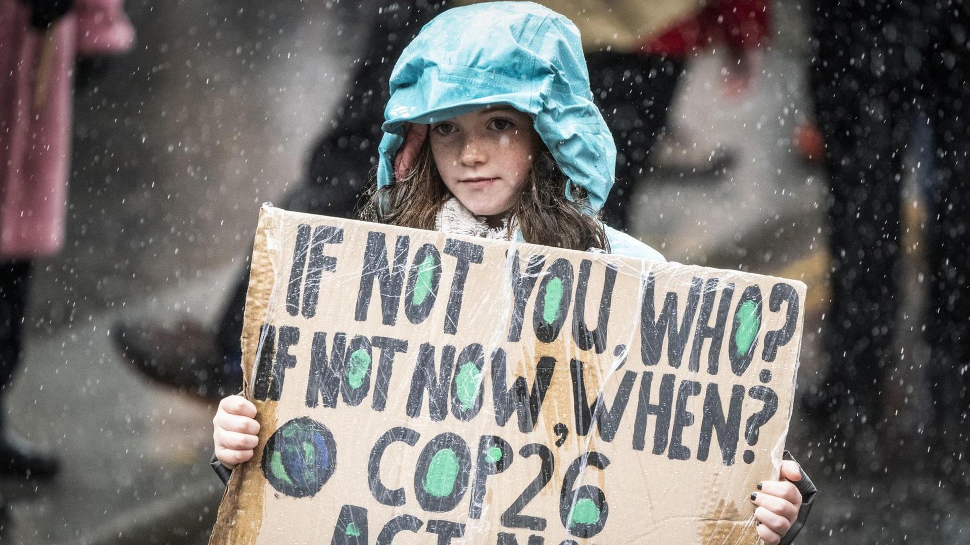 Ein junges Mädchen mit grüner Regenjacke trägt ein Pappschild, auf dem steht: "If not you, who? If not now, when? COP26 act now!! Es regnet.