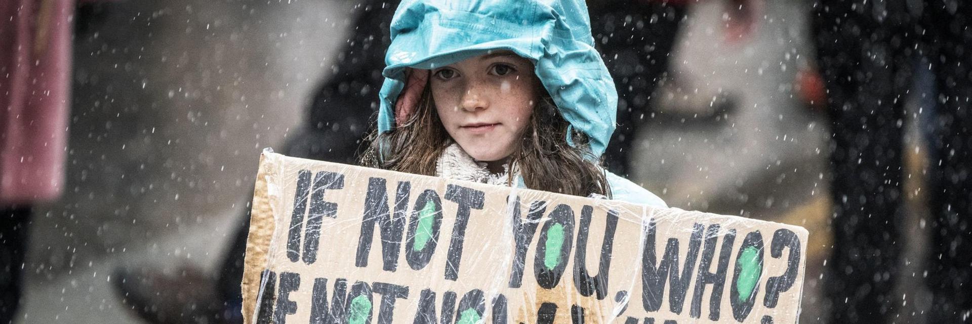 Ein junges Mädchen mit grüner Regenjacke trägt ein Pappschild, auf dem steht: "If not you, who? If not now, when? COP26 act now!! Es regnet.