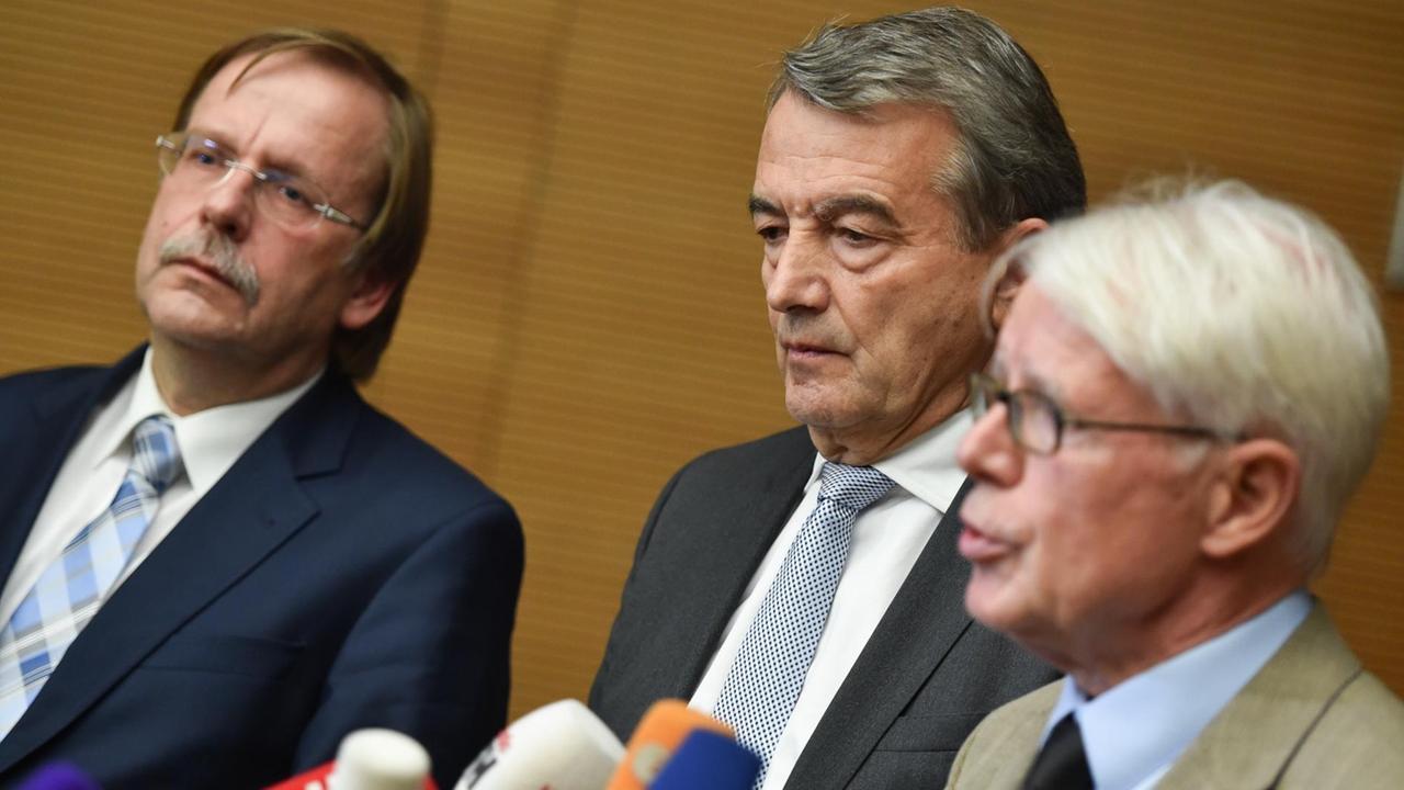 Die drei sitzen vor einer holzvertäfelten Wand. Rauball spricht in die zahlreichen Mikrofone.