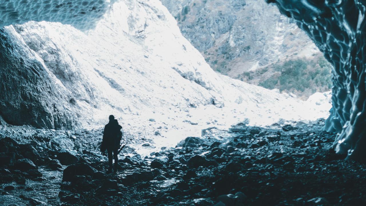 Ein Mann von hinten tritt aus einer Höhle und blickt auf schneebedecktes Gebirge. 