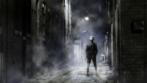 Ein Mann mit Hut und Anzug steht in nachts in einer spärlich beleuchteten Gasse.