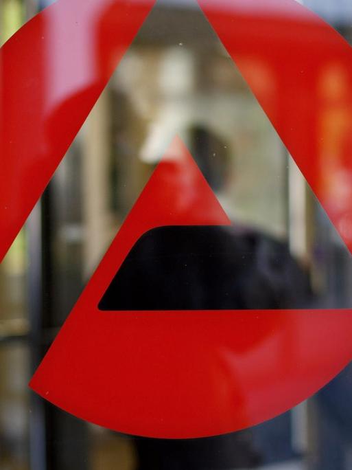 Eingang eines Jobcenters: Auf einer Scheibe klebt das rote Logo des Jobcenter, im Hintergrund in der Unschärfe betritt ein Mann das Gebäude.