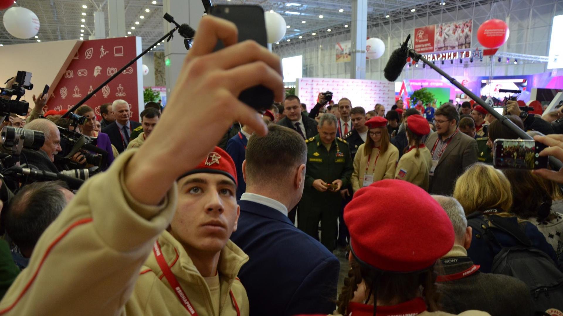 Ein Jungarmist in Uniform hält sein Smartphone in die Luft und macht ein Selfie. Im Hintergrund ist der Verteidigungsminister Sergej Schoigu zu sehen.