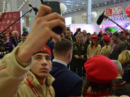 Ein Jungarmist in Uniform hält sein Smartphone in die Luft und macht ein Selfie. Im Hintergrund ist der Verteidigungsminister Sergej Schoigu zu sehen. Ein Jungarmist in Uniform hält sein Smartphone in die Luft und macht ein Selfie. Im Hintergrund ist der Verteidigungsminister Sergej Schoigu zu sehen.