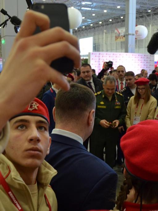 Ein Jungarmist in Uniform hält sein Smartphone in die Luft und macht ein Selfie. Im Hintergrund ist der Verteidigungsminister Sergej Schoigu zu sehen. Ein Jungarmist in Uniform hält sein Smartphone in die Luft und macht ein Selfie. Im Hintergrund ist der Verteidigungsminister Sergej Schoigu zu sehen.