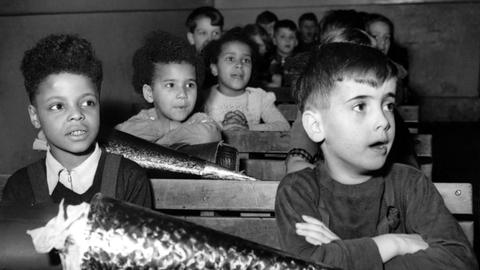 Drei farbige Besatzungskinder und Mitschüler an Tischen im Klassenzimmer, vor ihnen große, glitzernde Schultüten.
