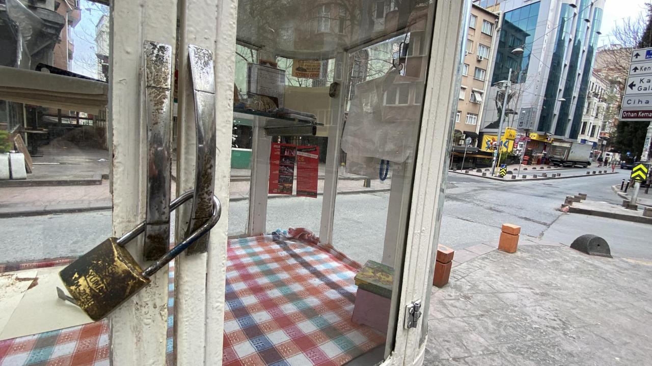 Im Vordergrund hält ein großes Vorhängschloß die weiße Tür eines kleinen Ladens mit karierter Tischdecke zusammen. Im Hintergrund erkennt man Straßenschilder, Straßen und Häuser.