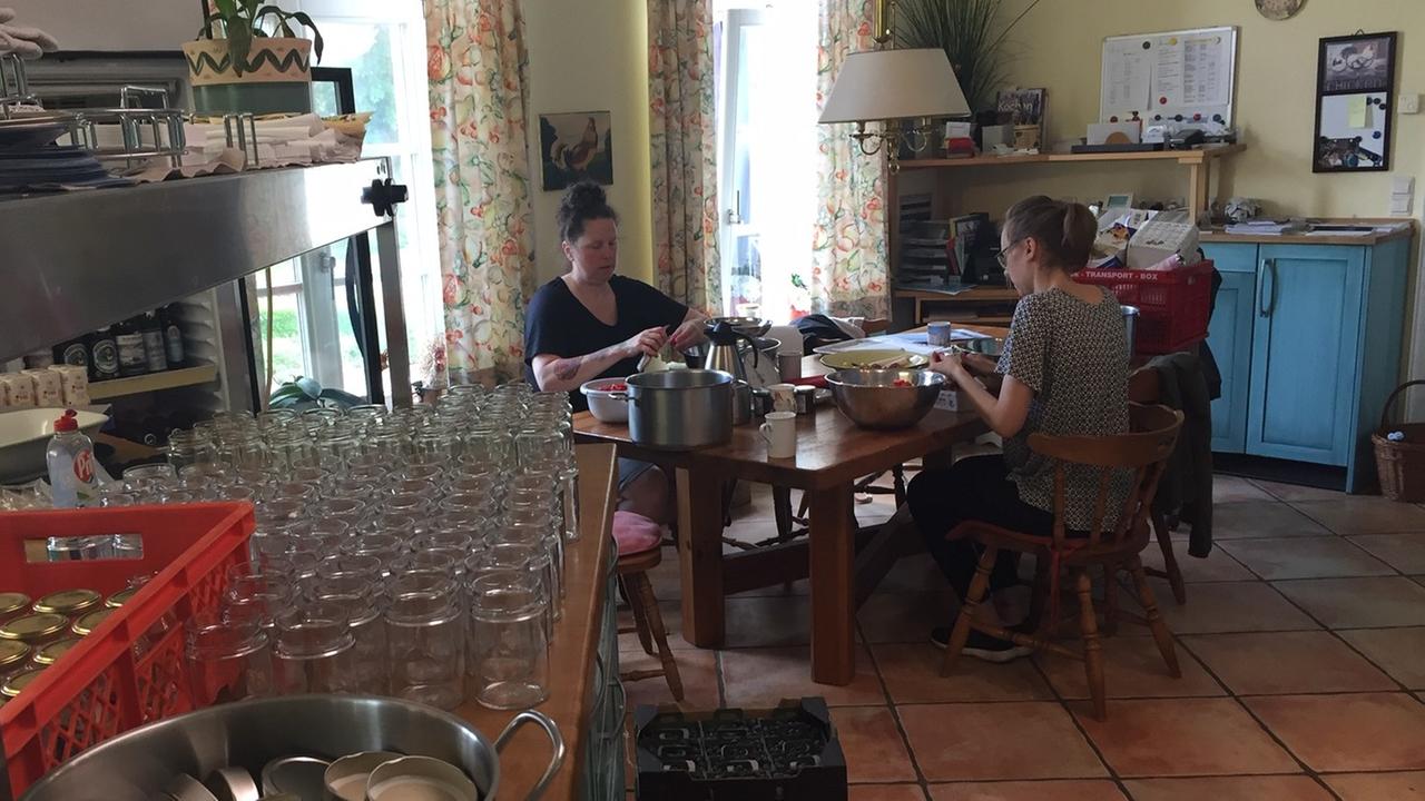 Zwei Frauen sitzen in einer Küche und putzen Früchte. Mehrere Kästen mit frischen Früchten stehen auf dem Boden der Küche. 