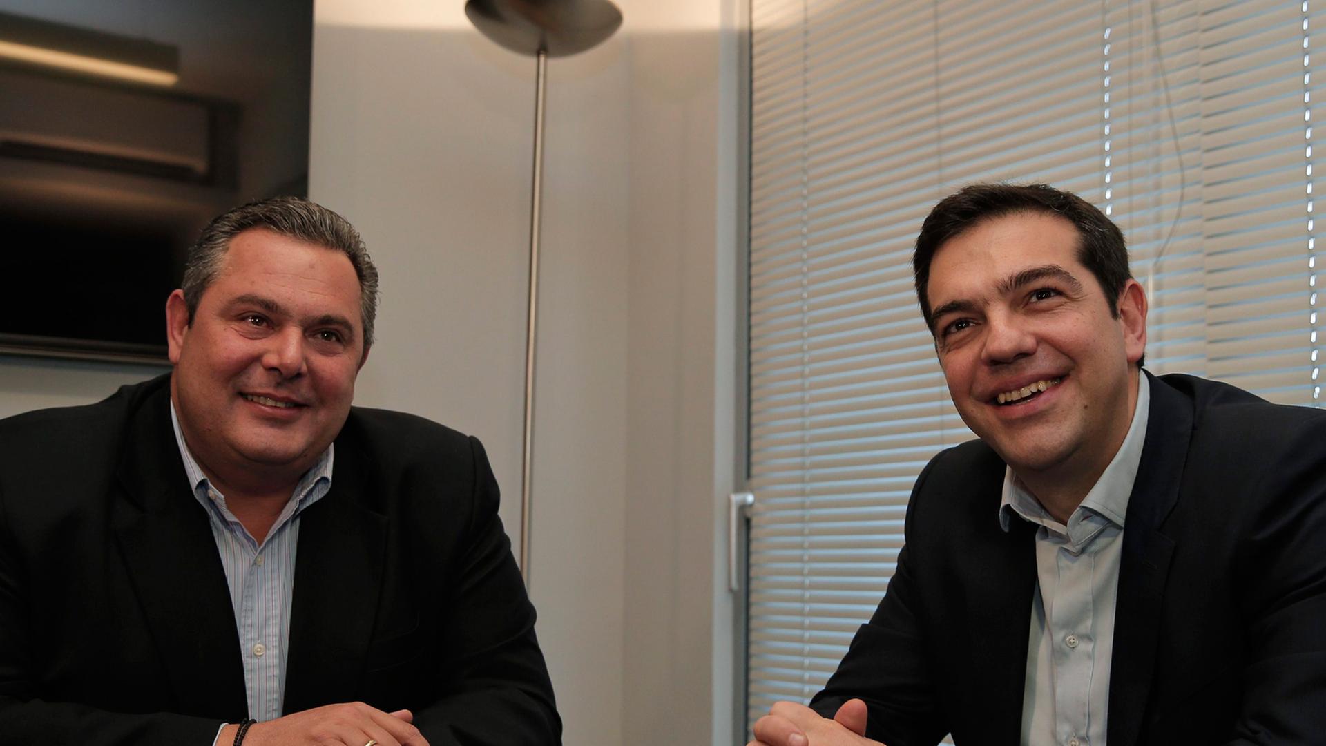 Panos Kammenos und Alexis Tsipras sitzen an einem Tisch und lächeln in die Kameras.