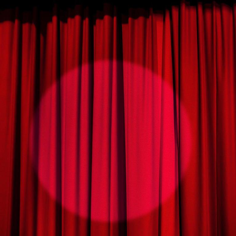Auf dem Foto sieht man einen Scheinwerfer, auf einen roten Theater-Vorhang gerichtet.
