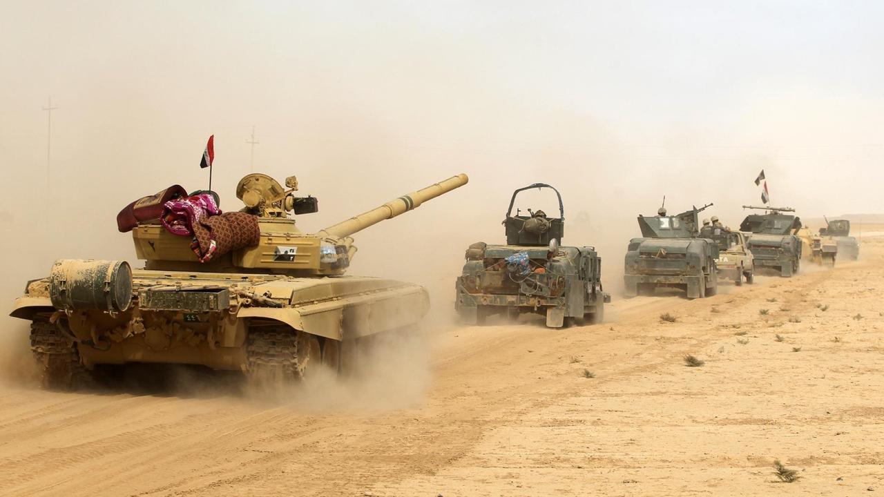 Panzer und Militärfahrzeuge rollen hintereinander durch den Wüstensand und ziehen Staubfahnenhinter sich her.