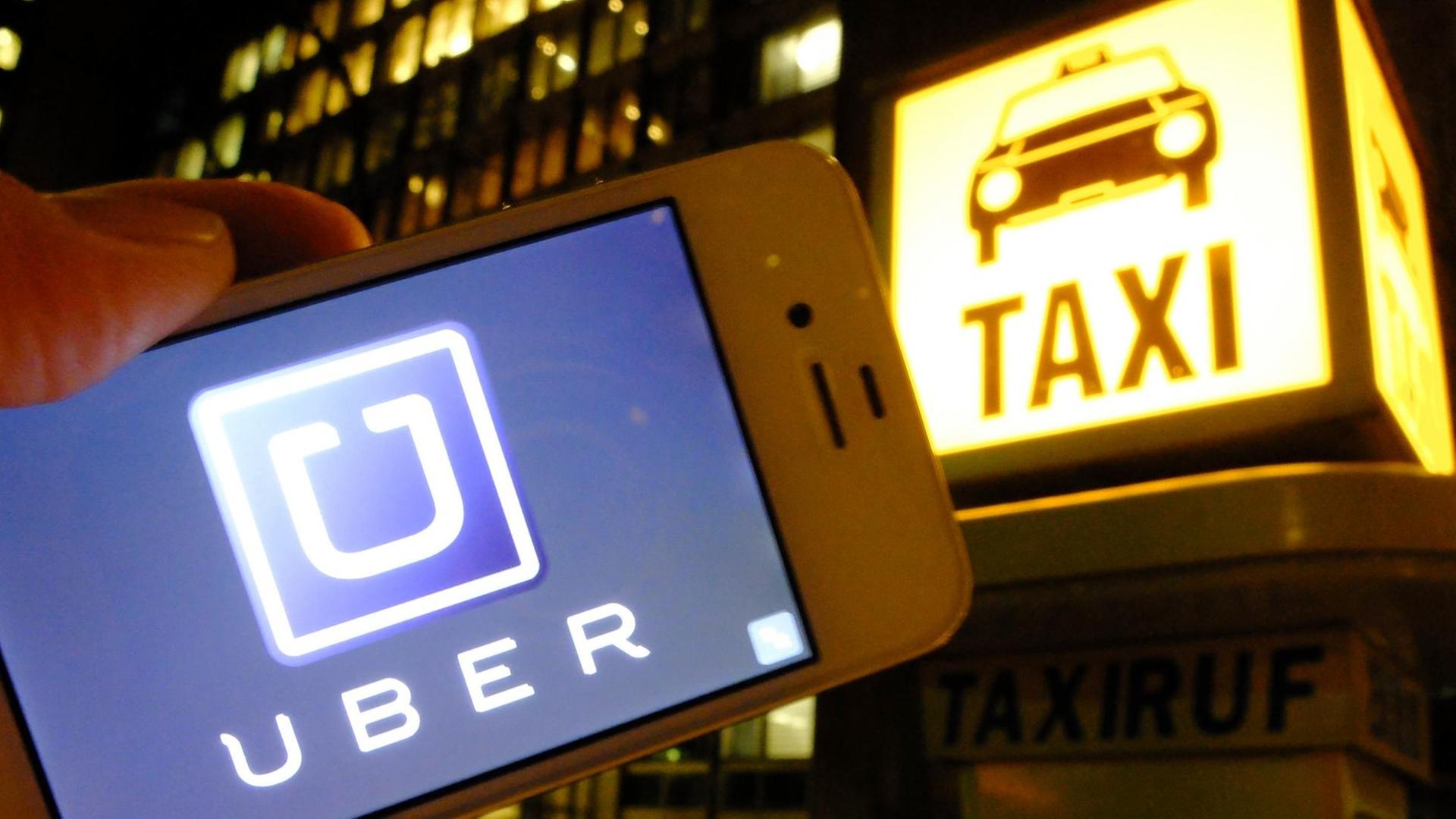 Es ist Nacht in einer Stadt: Eine Hand hält ein Smartphone mit dem Uber-Logo in die Kamera; im Hintergrund ist eine Taxirufsäule mit dem beleuchteten "Taxi"-Logo zu sehen.