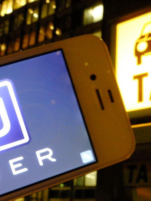 Es ist Nacht in einer Stadt: Eine Hand hält ein Smartphone mit dem Uber-Logo in die Kamera; im Hintergrund ist eine Taxirufsäule mit dem beleuchteten "Taxi"-Logo zu sehen.