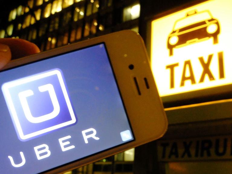Es ist Nacht in einer Stadt: Eine Hand hält ein Smartphone mit dem Uber-Logo in die Kamera; im Hintergrund ist eine Taxirufsäule mit dem beleuchteten "Taxi"-Logo zu sehen.