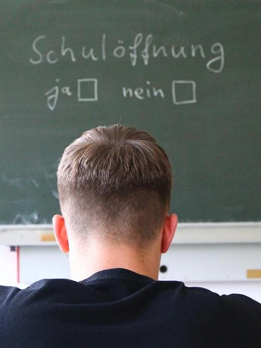 Ein Schüler sitzt vor einer Tafel, auf der "Schuloeffnung ja - nein" geschrieben steht.