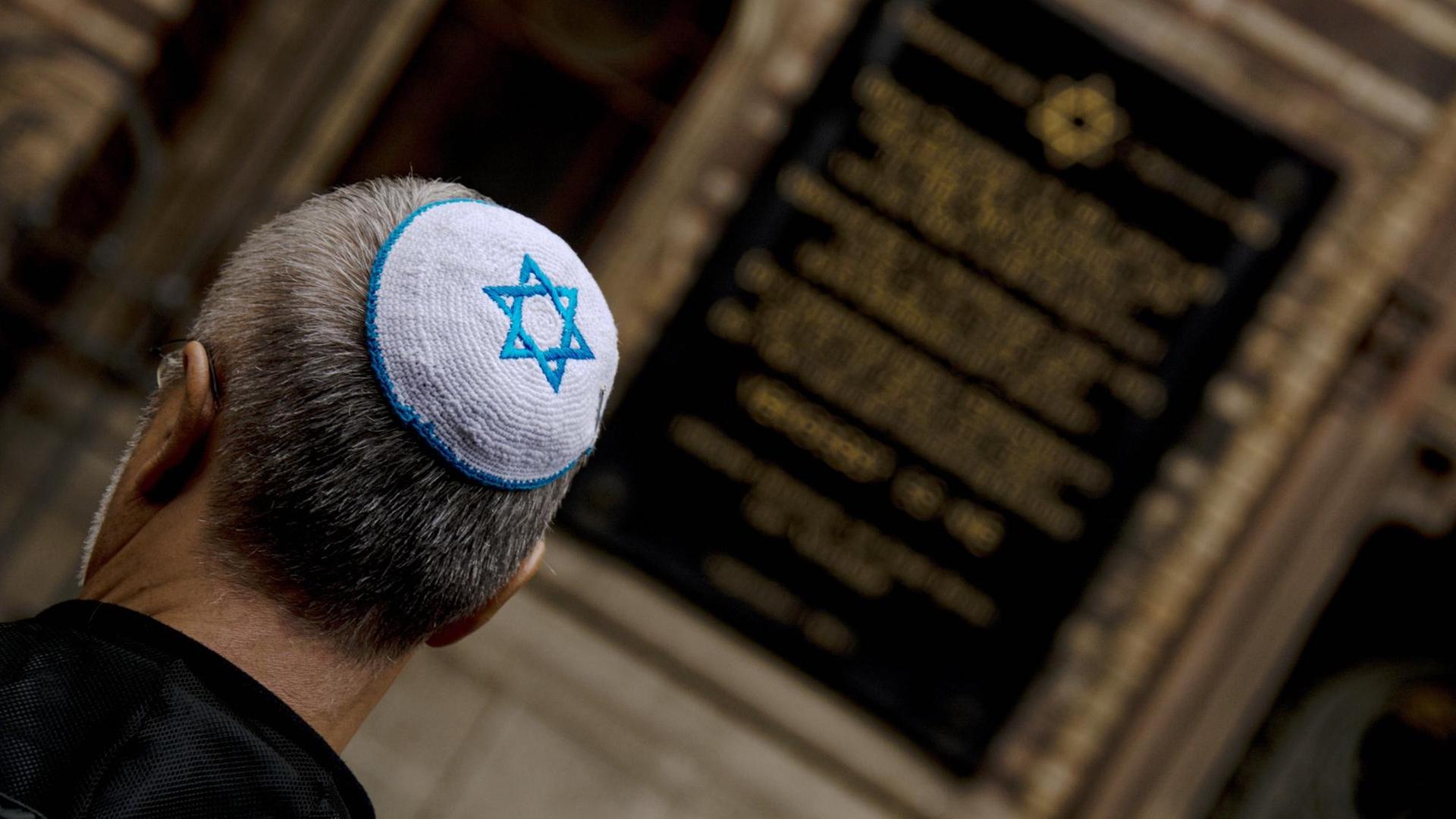 Rückansicht eines jüdischen Mannes mit Kippa, auf der sich ein blauer Judenstern auf weißem Grund befindet. Im Hintergrund zu sehen ist eine Fassadeninschrift.