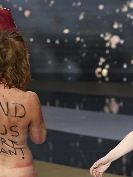 Die Schauspielerin Corinne Masiero steht nackt mit dem Rücken zur Kamera auf der Bühne bei der Verleihung des César. Auf Französisch steht auf ihrem Rücken "Gib uns die Kunst zurück, Jean!" Rechts von ihr steht angezogen die Moderatorin.