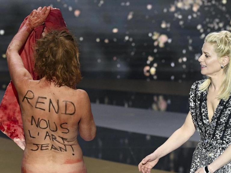 Die Schauspielerin Corinne Masiero steht nackt mit dem Rücken zur Kamera auf der Bühne bei der Verleihung des César. Auf Französisch steht auf ihrem Rücken "Gib uns die Kunst zurück, Jean!" Rechts von ihr steht angezogen die Moderatorin.