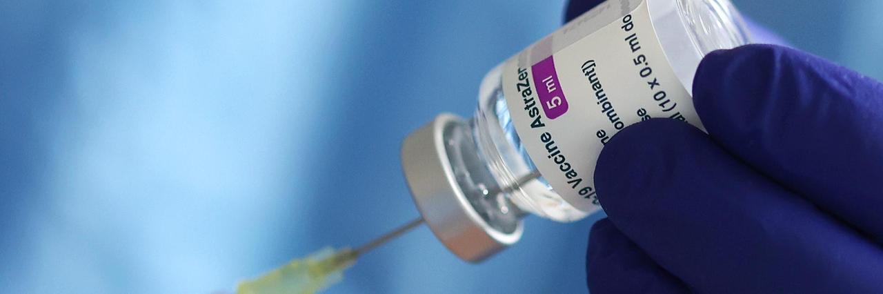 Ein blau behandschuhte Hand hält ein Fläschchen mit dem Covid-Impfstoff von AstraZeneca und präpariert eine Spritze. 