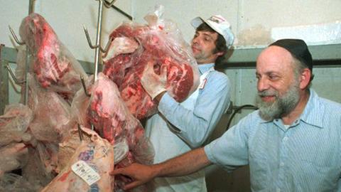Links im Bild ist geschächtetes Fleisch, rechts ein Metzger mit einer weißen Kappe und ein Mann mit einer schwarzen Kipa zu sehen.