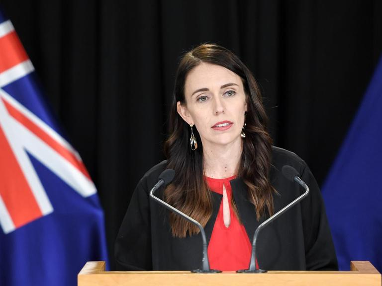 Porträt der neuseeländischen Premierministerin Jacinda Ardern am Rednerpult, umsäumt von Flaggen.