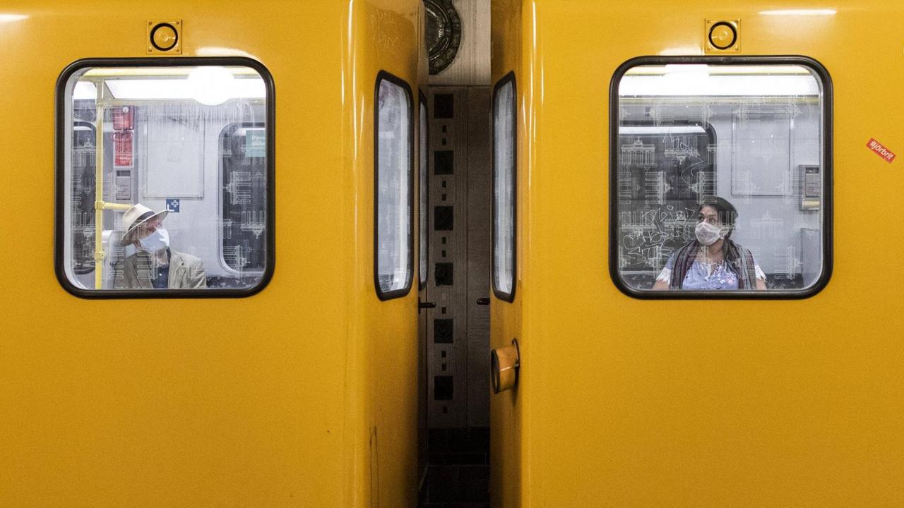 Zwei Personen mit Mund-Nasen-Maske sitzen in unterschiedlichen Waggons der Berliner U-Bahn.