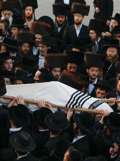 Zahlreiche orthodoxe Juden beobachten, wie ein eingehüllter Toter in einen Wagen getragen wird.