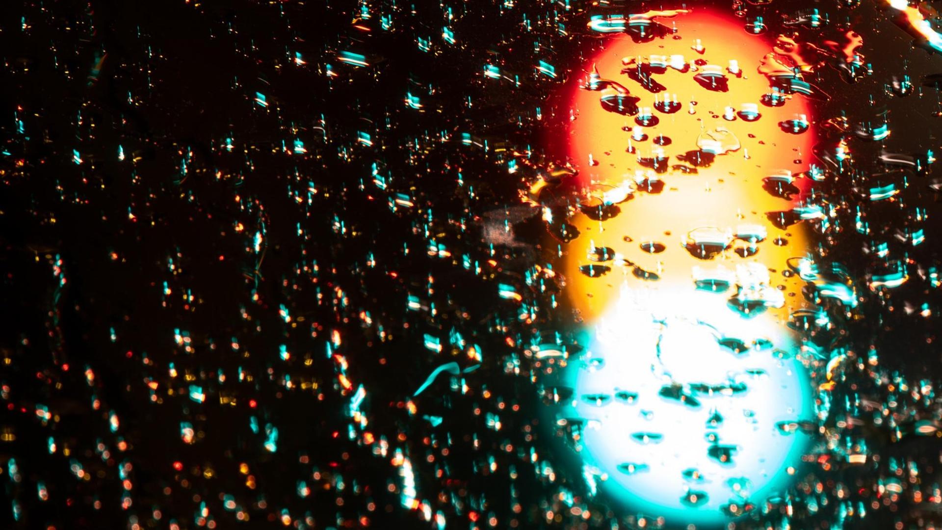 Eine Ampel leuchtet in allen Phasen Rot, Gelb und Grün. Über dem durch Langzeitbelichtung entstandenen Foto liegen Regentropfen.