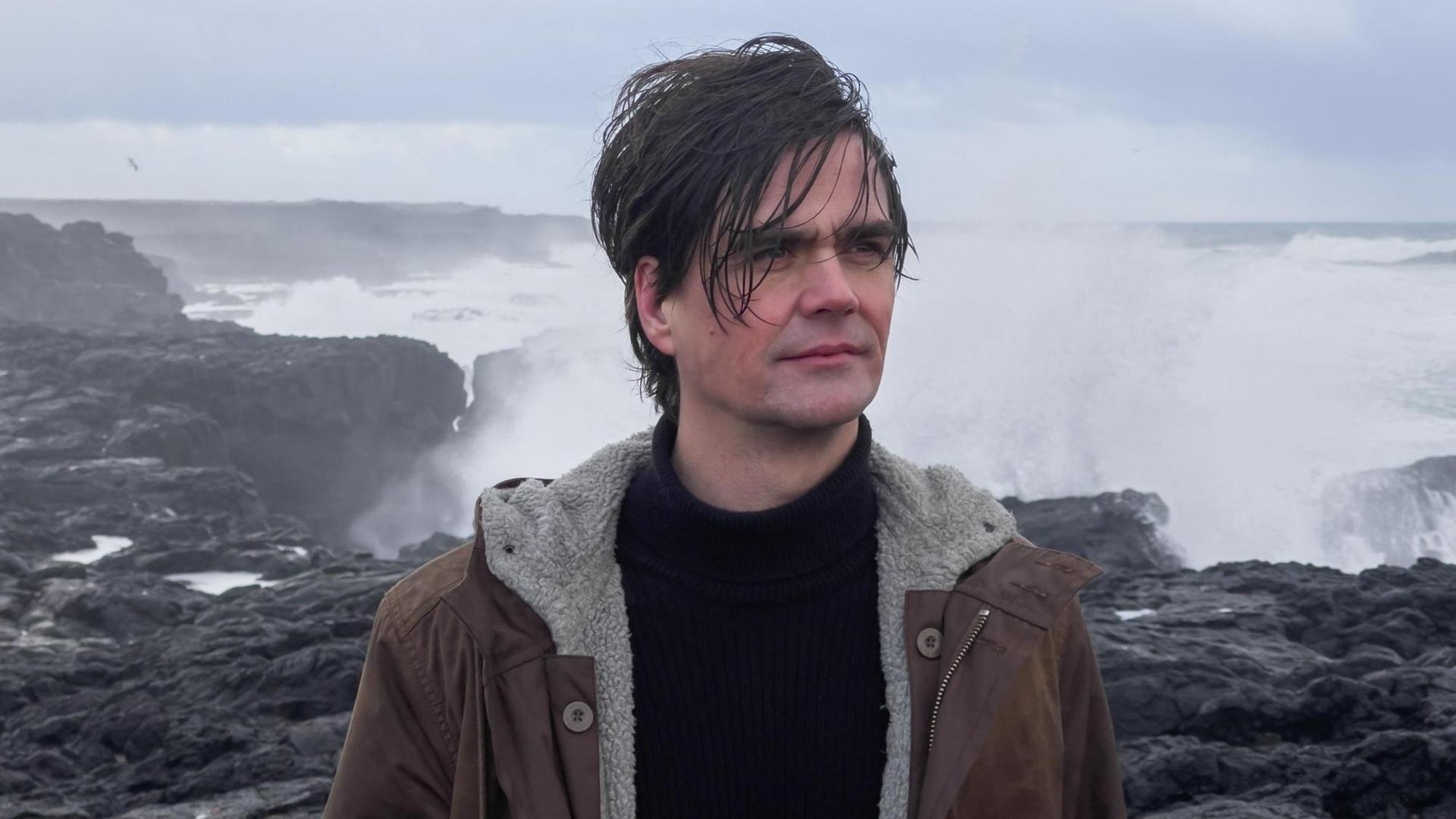 Ein Mann mit aufgewühlten Haar steht an einer Klippe vor tosendem Meer und schaut mit ernsthaften Blick in die Weite.