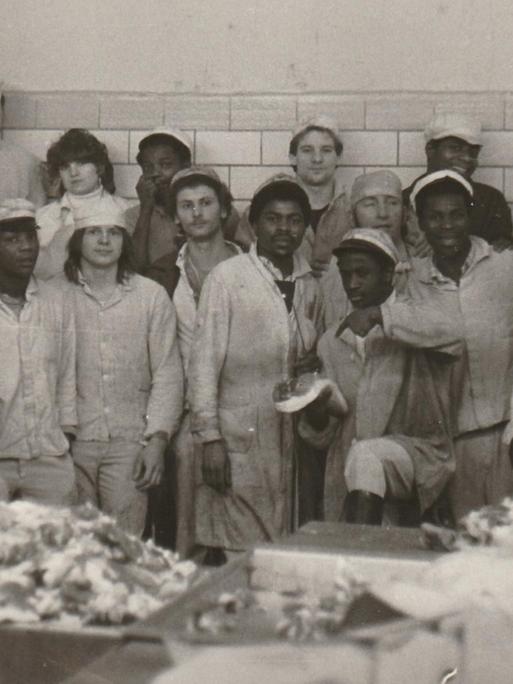 Gruppenfoto mit DDR-Vertragsarbeitern aus Mosambik im VEB Fleischkombinat Berlin 1982. Männer und Frauen in Arbeitskleidung posieren für ein Gruppenbild in einem fleischverarbeitenden Betrieb.