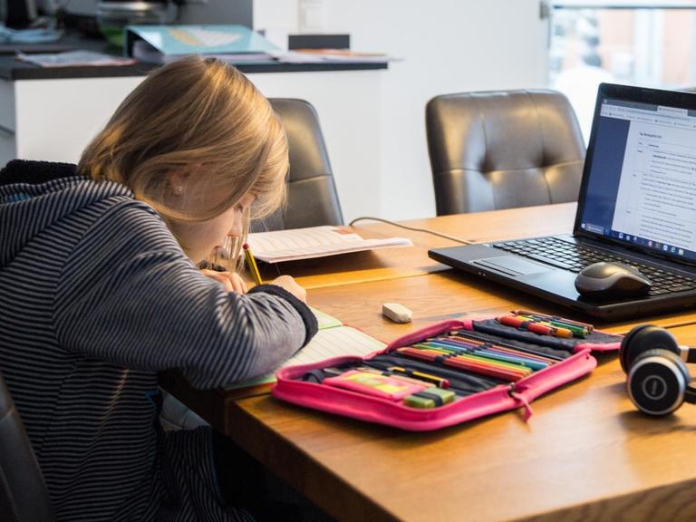Ein Kind sitzt an einem Küchentisch und löst Aufgaben in einem Schulheft. Auf dem Tisch steht ein aufgeklappter Laptop, neben dem Rechner liegen Bücher und Hefte mit weiteren Schulutensilien für das Homeschooling.