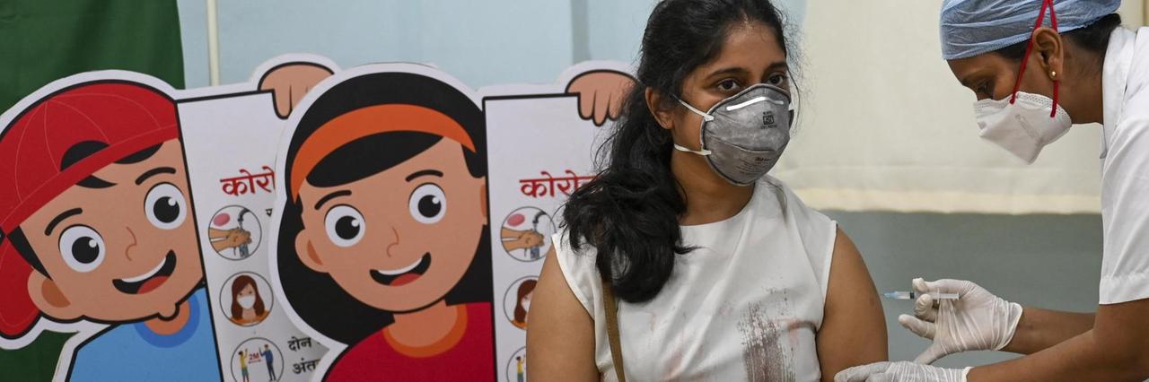Eine jüngere Frau wird in einem indischen Krankenhaus geimpft, hinter ihr stehen Schautafeln mit Hygieneregeln. 