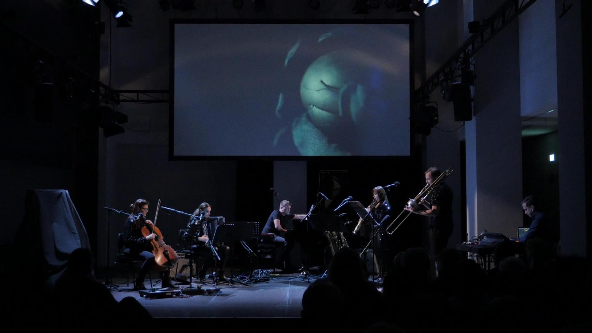 Sechs Musikerinnen und Musiker performen auf einer dunkelblau ausgeleuchteten Bühne. Über Ihnen im hängt eine Leinwand mit einer Projektion. Sechs Musikerinnen und Musiker performen auf einer dunkelblau ausgeleuchteten Bühne. Über Ihnen im hängt eine Leinwand mit einer Projektion.