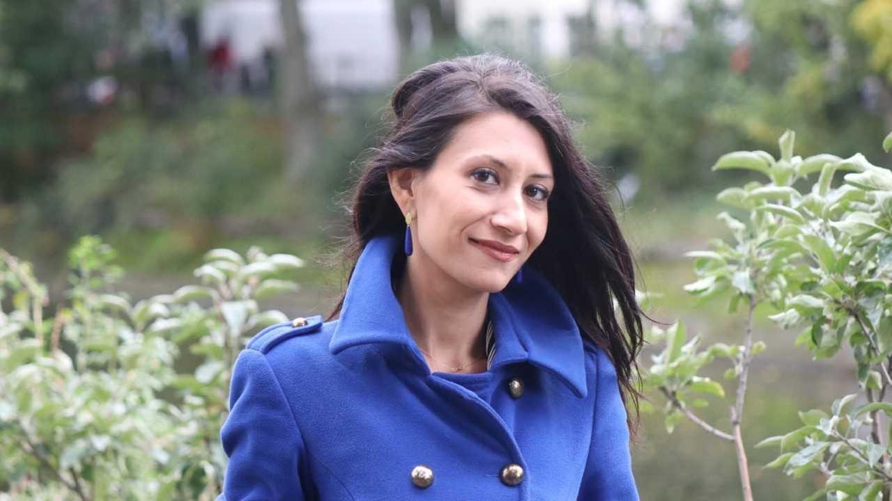 Eine schmale Frau mit dunklen langen Haaren in einem indigo-blauen Mantel. Der Kragen ist hochgeklappt. Sie steht in einem Park.
