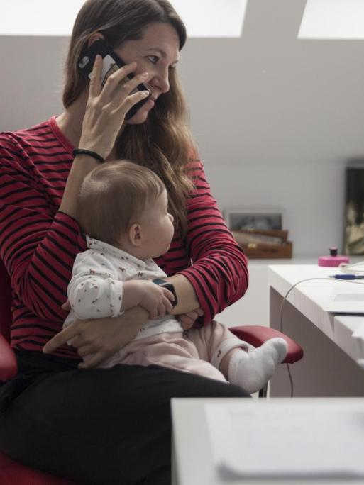 Eine Frau sitzt telefonierend am Schreibtisch, auf dem Schoß ihr Baby