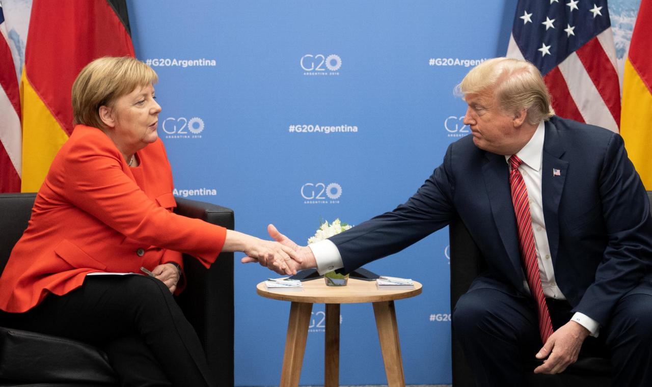 Das Bild zeigt Bundeskanzlerin Angela Merkel (CDU) und Donald Trump, Präsident der Vereinigten Staaten, Sie sitzen nebeneinander an einem Tisch und geben sich die Hand. Im Hinergrund sind die Flaggen Deutschlands und der USA sowie eine blaue Wand mit dem offiziellen Logo des Gipfeltreffens zu sehen. 