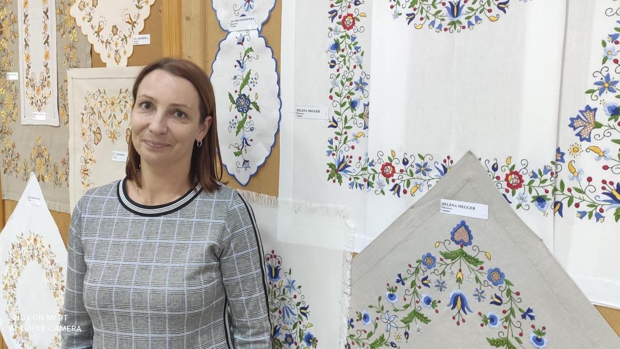 Kamila Soroko, die Kulturhausleiterin, steht im grau karierten Pullover vor einer Wand, an der unterschiedliche traditionelle Stickereien mit Blumenmustern aufgehängt sind.